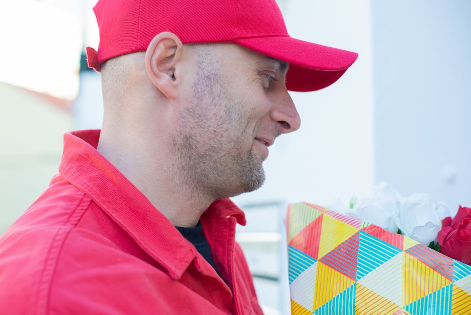 A delivery person dressed in a red jacket and matching red cap is holding a colorful gift box with a geometric pattern of red, yellow, blue, and white triangles, containing white and red flowers. The 