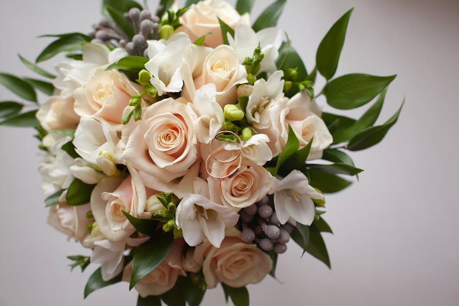A close-up of a floral bouquet featuring blush pink roses, white freesias, and small purple hypericum berries, accented with lush green foliage and wrapped with a clear glass vase. The flowers are fre
