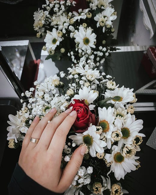 A hand with a gold wedding band gently touching a bouquet of fresh flowers, including a prominent red rose surrounded by white daisies with yellow centers and delicate baby's breath, arranged in a com