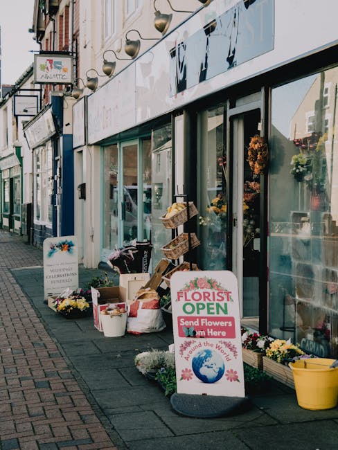 The image depicts the exterior of a florist shop on Teddington High Street, with large glass windows revealing a display of vibrant flower arrangements inside. Outside, there are various fresh floral 