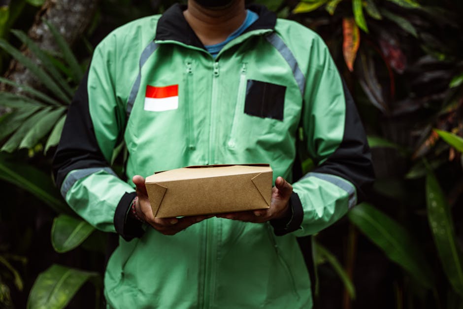 A delivery person wearing a bright green and black jacket with reflective stripes, standing outdoors against a backdrop of lush green foliage. The jacket features a national flag patch on the chest an